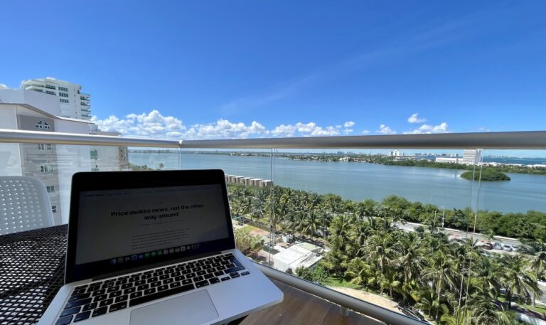 Laptop on a balcony table overlooking a lagoon, palm trees and hotel towers under a blue sky